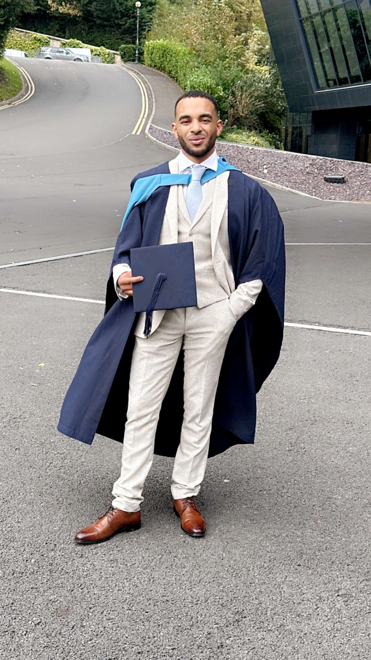 A young man stands outdoors wearing a light-colored suit, blue tie, and blue graduation gown, holding a graduation cap and certificate in one hand. He stands confidently on a paved road with greenery, trees, and a modern glass building in the background. The scene conveys a sense of pride and accomplishment.