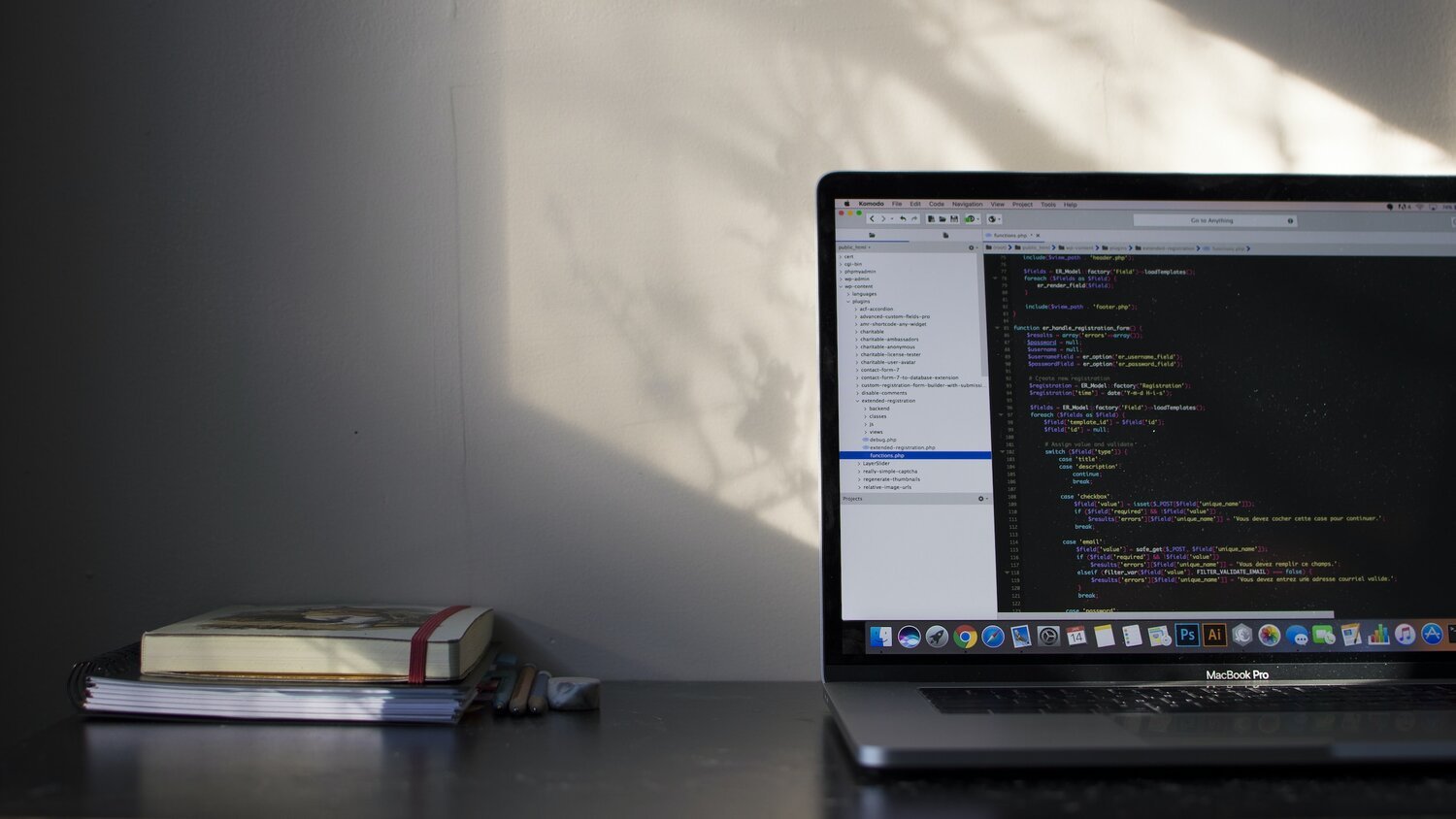 Open MacBook Pro displaying a code editor with colorful syntax highlighting, positioned on a dark desk next to a small stack of books and pencils. Soft natural light and plant shadows fall across a plain wall, creating a calm and focused workspace. No visible text in the environment except for code on the laptop screen.